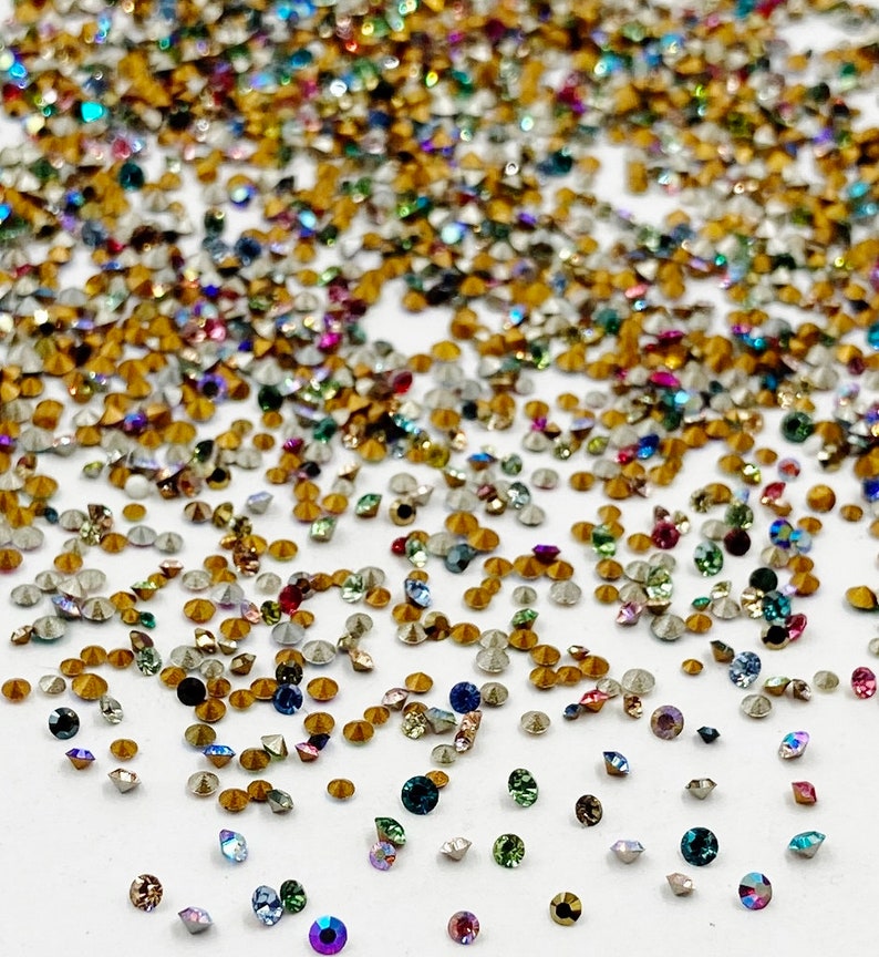 May include: A large assortment of colorful rhinestones, with a variety of shapes and sizes, scattered on a white surface.