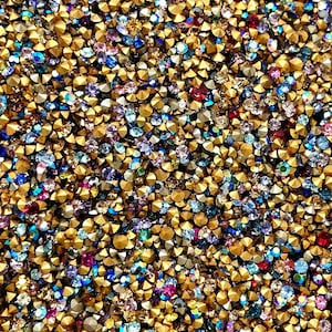 May include: A close-up image of a large quantity of small, colorful rhinestones. The rhinestones are mixed with gold-colored metal studs. The rhinestones are various shades of blue, purple, red, and green.