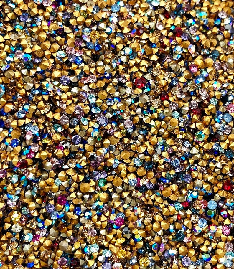 May include: A close-up image of a large quantity of small, colorful rhinestones. The rhinestones are mixed with gold-colored metal studs. The rhinestones are various shades of blue, purple, red, and green.