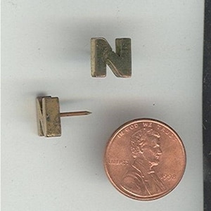 May include: Two gold-colored letter N pins. One pin is attached to a push pin. The other pin is laying flat on a white surface. A penny is in the background.