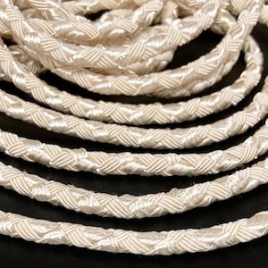 May include: A continuous length of off-white braided cord featuring a dense, woven texture with a subtle sheen. The cord is coiled in multiple overlapping circular layers, showcasing the intricate, repeating diagonal pattern of the braided strands.