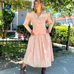 May include: A light pink vintage dress with a white lace collar and gold embellishments. The dress has a fitted bodice and a full skirt.