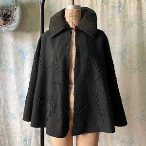 May include: A black vintage cape with a wide collar and leaf-like embroidery. The cape is displayed on a mannequin, showcasing its flowing design and intricate details. The dark color and unique embellishments suggest a historical or theatrical style.