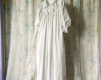May include: A long, white nightdress with a frilled neckline and cuffs hangs from a metal hanger. The nightdress has long sleeves and a full skirt. The background is a floral patterned curtain.