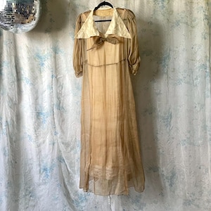 May include: A vintage, floor-length dress in shades of tan and cream, hanging on a black hanger. The dress features a cream-colored collar and a bow detail at the neckline. The sheer fabric creates a flowing silhouette, with a lace trim at the hem.