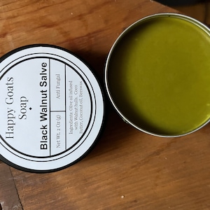 May include: A white tin container with a black label that reads "Happy Goats Soap Black Walnut Salve Net Wt. 2 Oz (g) Anti Fungal Ingredients: Olive oil infused with Walnut hulls. Coco Butter, Coconut oil, Beeswax" and a green salve in a tin container.