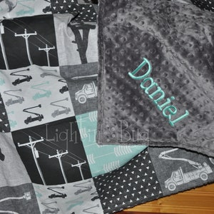May include: A gray and white baby blanket with a patchwork design featuring a variety of images, including trucks, power lines, and geometric patterns. The blanket is personalized with the name "Daniel" embroidered in teal on the gray side.