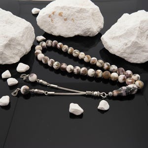 May include: A beaded necklace with a tassel, featuring round, speckled beads in shades of white, brown, and purple. The necklace has silver-coloured metal accents and is displayed on a black surface with white rocks.