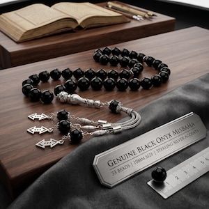 May include: A black onyx misbaha with sterling silver accents, featuring 33 beads, each 10mm in size. The prayer beads are displayed on a wooden surface alongside a ruler and an open book. The metal tag reads "GENUINE BLACK ONYX MISBAHA".