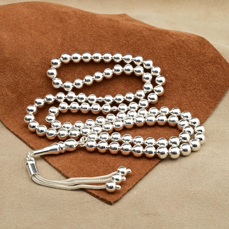 May include: A silver prayer beads necklace with round beads. The necklace is on a brown leather surface.