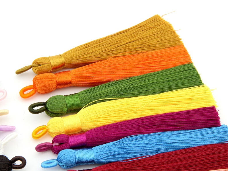 May include: Assortment of colorful silk tassels in shades of gold, orange, green, yellow, purple, blue, and red. The tassels are arranged in a row, with the gold tassel at the top and the red tassel at the bottom.
