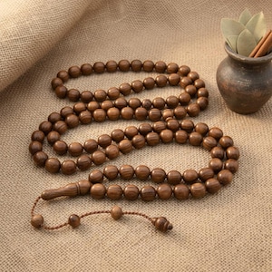 May include: A wooden prayer bead set, comprising numerous round beads strung together. The beads are a warm brown colour, with a smooth, polished finish. The set includes a tassel and decorative end pieces. A small, dark brown ceramic vase with leaves and cinnamon sticks is in the background.