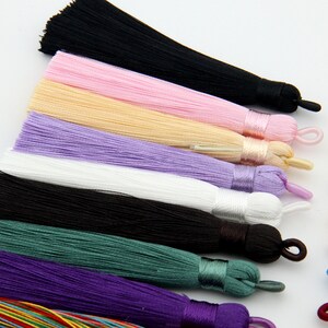 May include: Assortment of colorful silk tassels in various colors, including black, pink, beige, purple, white, green, and purple. The tassels are arranged in a row, with the black tassel at the top and the purple tassel at the bottom.