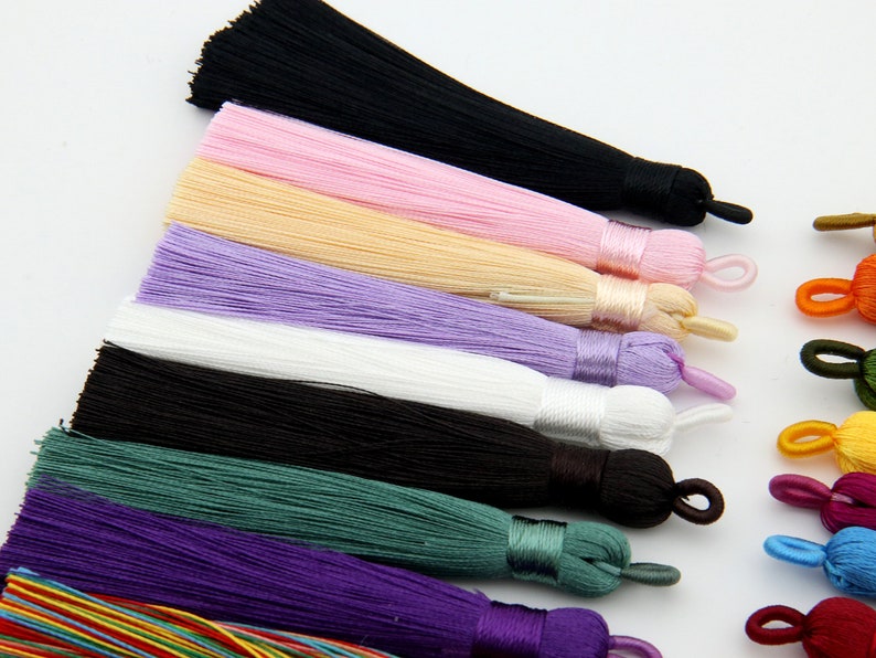 May include: Assortment of colorful silk tassels in various colors, including black, pink, beige, purple, white, green, and purple. The tassels are arranged in a row, with the black tassel at the top and the purple tassel at the bottom.