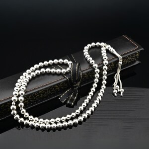 May include: A silver prayer beads necklace with a tassel, displayed in a black box with a gold ribbon.
