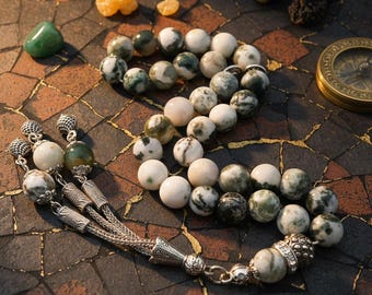 Può includere: Un rosario di preghiera con perline maculate bianche, verdi e grigie. Il rosario presenta decorazioni in metallo color argento nella parte superiore e inferiore, con un nappa. Una bussola e altri piccoli oggetti sono sullo sfondo.