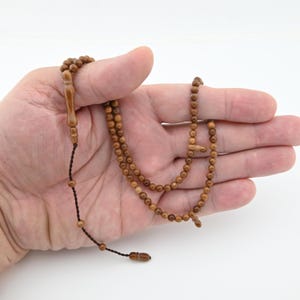 May include: A wooden prayer bead necklace with small, round beads in varying shades of brown. The beads are strung on a dark cord, with a decorative wooden piece at the top and a tassel at the end. The necklace is held in a hand.