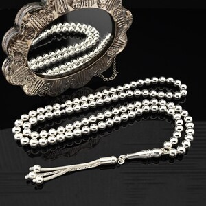 May include: A silver prayer beads necklace with a tassel. The beads are round and smooth. The necklace is laid out on a black surface.
