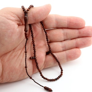 May include: A dark brown prayer bead necklace with small, round beads and a decorative elongated bead. The necklace is held in a hand against a white background. The beads are strung on a dark brown cord.