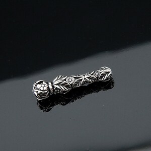 May include: A silver-colored, cylindrical bead with intricate floral and leaf patterns. The bead has a rounded end with a flower design and is approximately 1.5 inches long. The detailed carvings give it a vintage or handcrafted appearance.