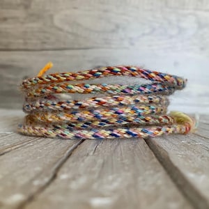 May include: A close-up of a multi-coloured braided cord with a variety of colours including blue, red, yellow, and green. The cord is coiled on a wooden surface.