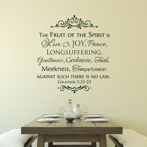 May include: A wall decal with a green, cursive font that reads "The Fruit of the Spirit is Love, Joy, Peace, Longsuffering, Gentleness, Goodness, Faith, Meekness, Temperance: Against such there is no law. Galatians 5:22-23". The decal is on a beige wall above a wooden table with a white tablecloth and a glass pitcher and glasses.