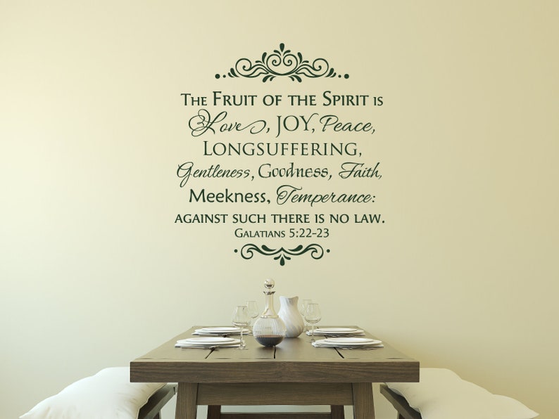 May include: A wall decal with a green, cursive font that reads "The Fruit of the Spirit is Love, Joy, Peace, Longsuffering, Gentleness, Goodness, Faith, Meekness, Temperance: Against such there is no law. Galatians 5:22-23". The decal is on a beige wall above a wooden table with a white tablecloth and a glass pitcher and glasses.