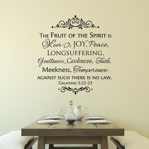 May include: A black and white wall decal with a quote from the Bible: "The fruit of the Spirit is love, joy, peace, longsuffering, gentleness, goodness, faith, meekness, temperance: against such there is no law. Galatians 5:22-23". The decal is on a beige wall above a wooden table with a vase and plates.