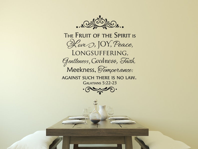May include: A black and white wall decal with a quote from the Bible: "The fruit of the Spirit is love, joy, peace, longsuffering, gentleness, goodness, faith, meekness, temperance: against such there is no law. Galatians 5:22-23". The decal is on a beige wall above a wooden table with a vase and plates.