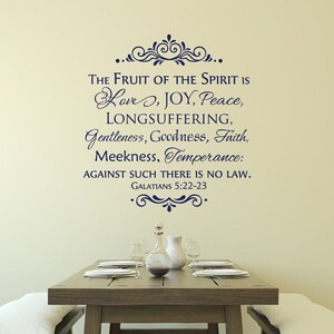 May include: A wall decal with a navy blue script quote from the Bible, "The fruit of the Spirit is love, joy, peace, longsuffering, gentleness, goodness, faith, meekness, temperance: against such there is no law. Galatians 5:22-23". The decal is displayed on a beige wall above a wooden dining table with a white tablecloth and a glass pitcher.