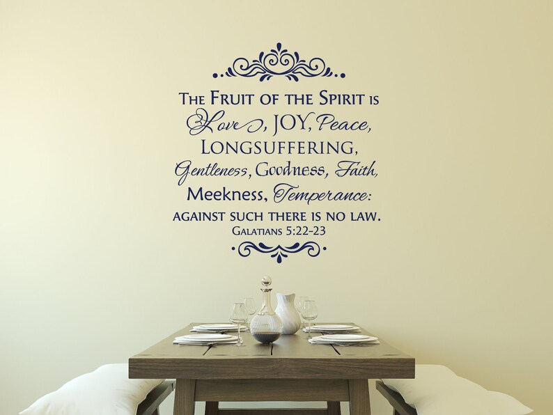 May include: A wall decal with a navy blue script quote from the Bible, "The fruit of the Spirit is love, joy, peace, longsuffering, gentleness, goodness, faith, meekness, temperance: against such there is no law. Galatians 5:22-23". The decal is displayed on a beige wall above a wooden dining table with a white tablecloth and a glass pitcher.