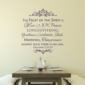 May include: A wall decal with a quote from the Bible, Galatians 5:22-23, that reads "The fruit of the Spirit is love, joy, peace, longsuffering, gentleness, goodness, faith, meekness, temperance: against such there is no law." The decal is in a dark purple color and is placed on a light beige wall above a wooden table with a vase and plates.