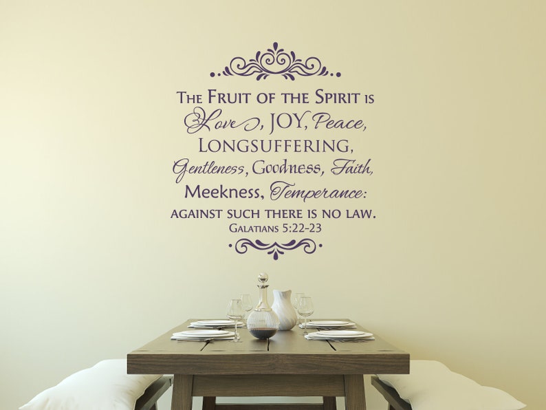 May include: A wall decal with a quote from the Bible, Galatians 5:22-23, that reads "The fruit of the Spirit is love, joy, peace, longsuffering, gentleness, goodness, faith, meekness, temperance: against such there is no law." The decal is in a dark purple color and is placed on a light beige wall above a wooden table with a vase and plates.