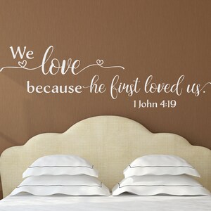 May include: White vinyl wall decal with the text "We love because he first loved us. 1 John 4:19" on a brown wall above a beige headboard with white pillows.