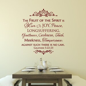 May include: A wall decal with a burgundy-colored quote from the Bible, "The fruit of the Spirit is love, joy, peace, longsuffering, gentleness, goodness, faith, meekness, temperance: against such there is no law. Galatians 5:22-23." The decal is on a beige wall above a wooden table set with white plates and a glass pitcher.