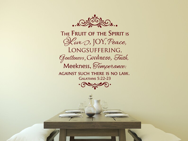 May include: A wall decal with a burgundy-colored quote from the Bible, "The fruit of the Spirit is love, joy, peace, longsuffering, gentleness, goodness, faith, meekness, temperance: against such there is no law. Galatians 5:22-23." The decal is on a beige wall above a wooden table set with white plates and a glass pitcher.