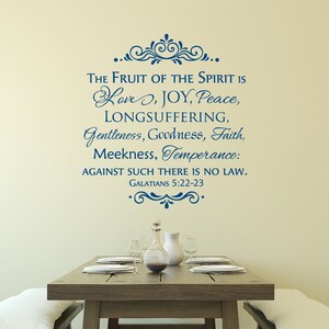 May include: A wall decal with a blue script quote from the Bible, "The Fruit of the Spirit is Love, Joy, Peace, Longsuffering, Gentleness, Goodness, Faith, Meekness, Temperance: Against such there is no law. Galatians 5:22-23". The decal is on a beige wall above a wooden table set with a vase and plates.