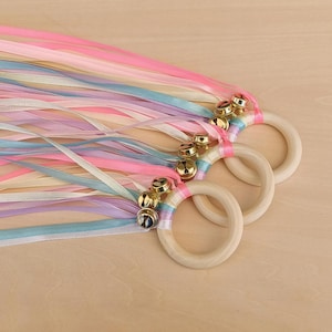 May include: Three wooden rings, each featuring a cluster of small gold-toned metal bells attached to the top. Multiple long, thin satin ribbons in shades of light pink, pale blue, white, and lavender are tied to each ring, flowing outward.