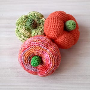 May include: Three crocheted pumpkins in different colors: green and yellow, orange, and pink and orange. The pumpkins are all about the same size and have a small green stem on top.