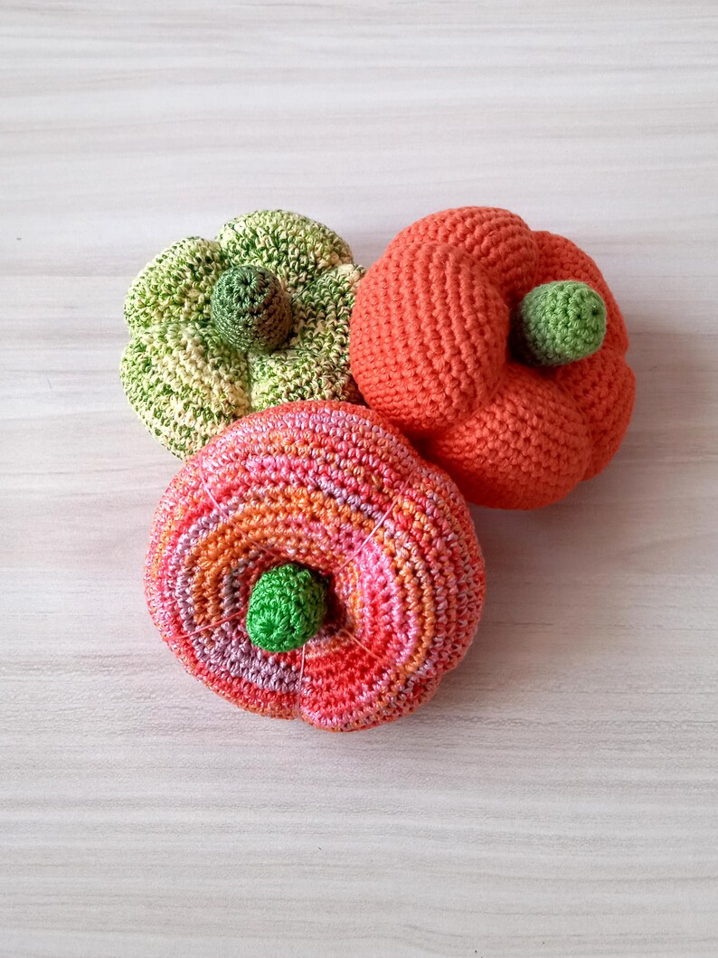 May include: Three crocheted pumpkins in different colors: green and yellow, orange, and pink and orange. The pumpkins are all about the same size and have a small green stem on top.