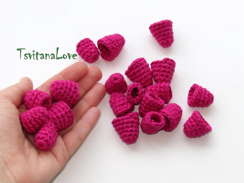 May include: A hand holding a pile of small, pink crocheted flower buds. There are more flower buds scattered on a white surface.