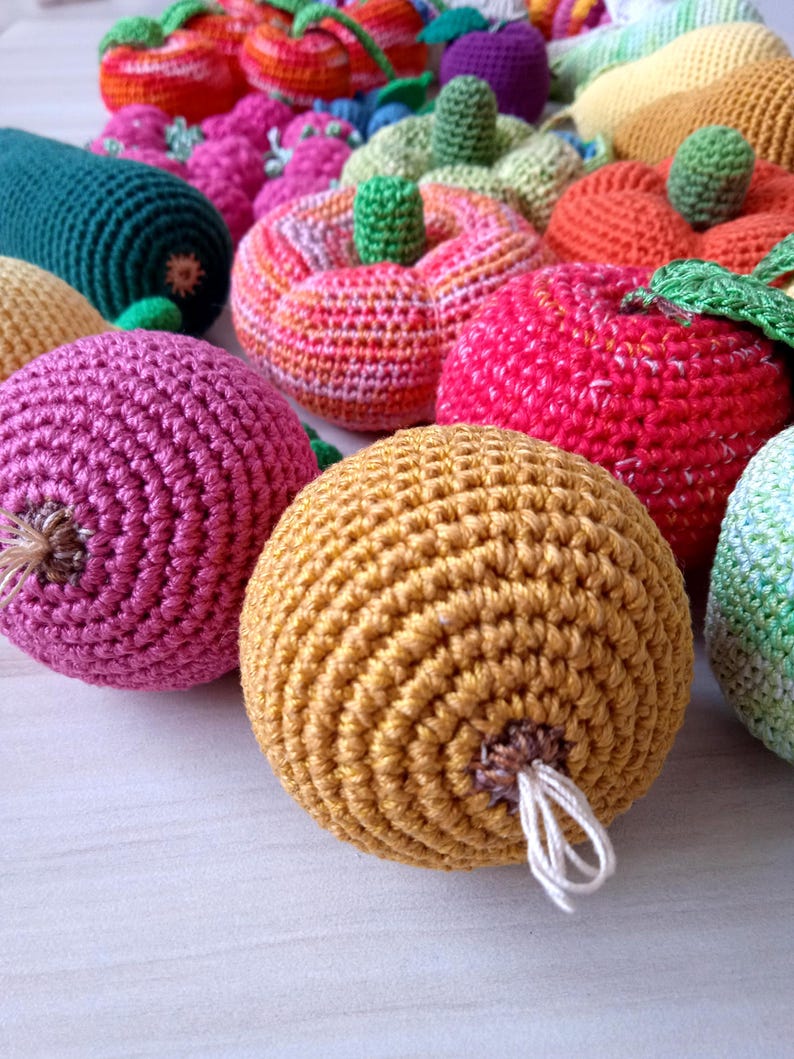 May include: A collection of crocheted fruits and vegetables, including a green zucchini, a pink beet, a yellow onion, a red apple, a green and orange pumpkin, and an orange tomato.