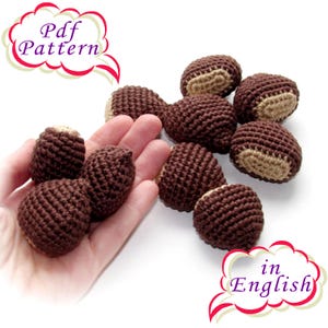 Op de afbeelding: Een hand houdt een groep van bruine, gehaakte kastanjevormige ornamenten vast. De ornamenten hebben een beige gehaakt detail aan één kant. De tekst "Pdf Pattern in English" staat in een spreekballon in de rechterbenedenhoek van de afbeelding.