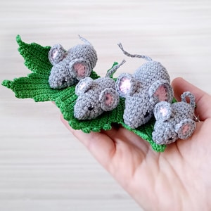 May include: Four gray crocheted mice with pink ears are sitting on a green crocheted leaf.