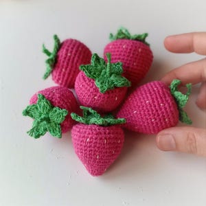 Könnte beinhalten: Sechs gehäkelte Erdbeeren in Pink mit grünen Blättern. Die Erdbeeren sind in einer Gruppe auf einer weißen Oberfläche angeordnet.
