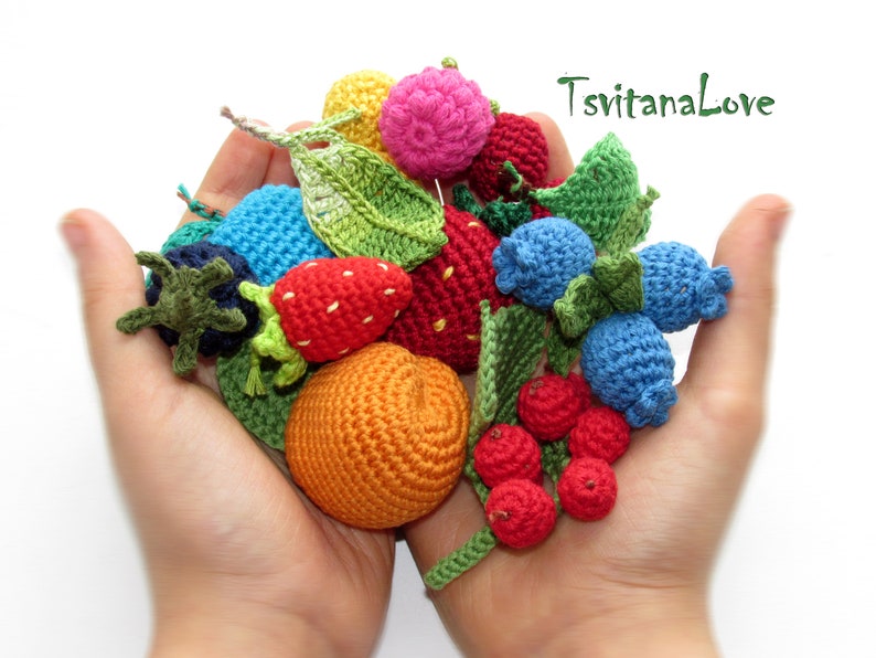 May include: A collection of crocheted fruits and berries, including strawberries, blueberries, raspberries, and an orange, in a variety of colors. The crocheted fruits are arranged in a person's hands.