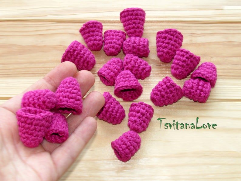 May include: A collection of 17 crocheted raspberry-shaped ornaments in a vibrant pink color. The ornaments are arranged on a wooden surface and in a hand.