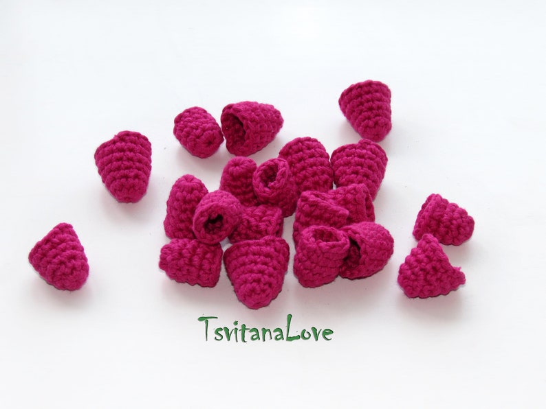 May include: A collection of 15 crocheted raspberry shapes in a vibrant pink color. The raspberries are arranged in a cluster on a white surface.