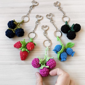 Pode incluir: Uma coleção de chaveiros de crochê feitos à mão com várias frutas. Os chaveiros incluem amoras, morangos, framboesas e mirtilos, cada um com folhas verdes e fechos prateados em forma de coração. As frutas são em tons de vermelho, rosa, azul e preto.