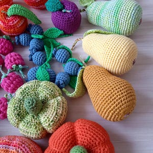 May include: A collection of crocheted fruit and vegetables, including a red apple, a purple plum, a green pear, a yellow pear, a green zucchini, a red pumpkin, and a cluster of blueberries.